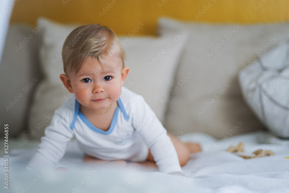 cute happy one year old happy blond babyboy in a white body or suit at home on a cozy bed after bathing and looking into the Camera 