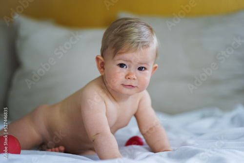 super sweet naked almost one year old happy blond baby boy lying at home on a cozy bed after bathing in prone position and smiling or flirting with the Camera like a innocent rascal