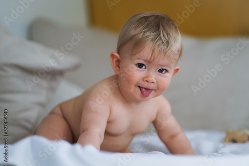 super sweet naked almost one year old happy blond baby boy lying at home on a cozy bed after bathing in prone position and smiling or flirting with the Camera like a innocent rascal