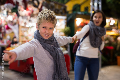 Preteen boy pulling his mom hand, demanding to buy something for him at outdoor Christmas market © JackF