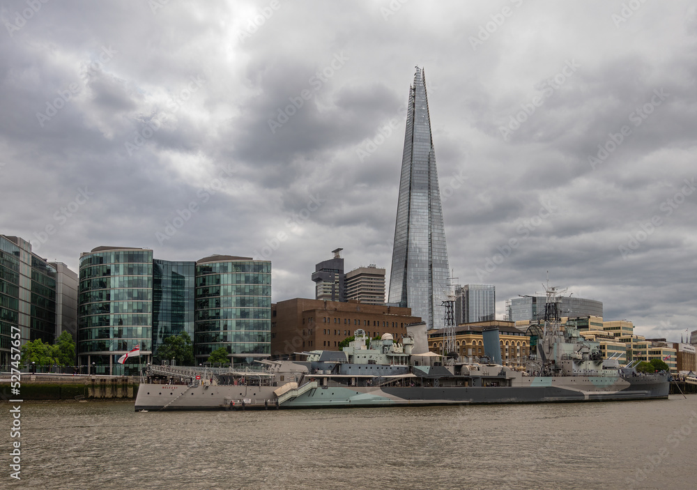 London, England, UK - July 6, 2022: From Thames River. Closeup, HMS Belfast museum ship plus The Chard skyscraper in back under gray cloudscape. Other buildings along Queens Walk