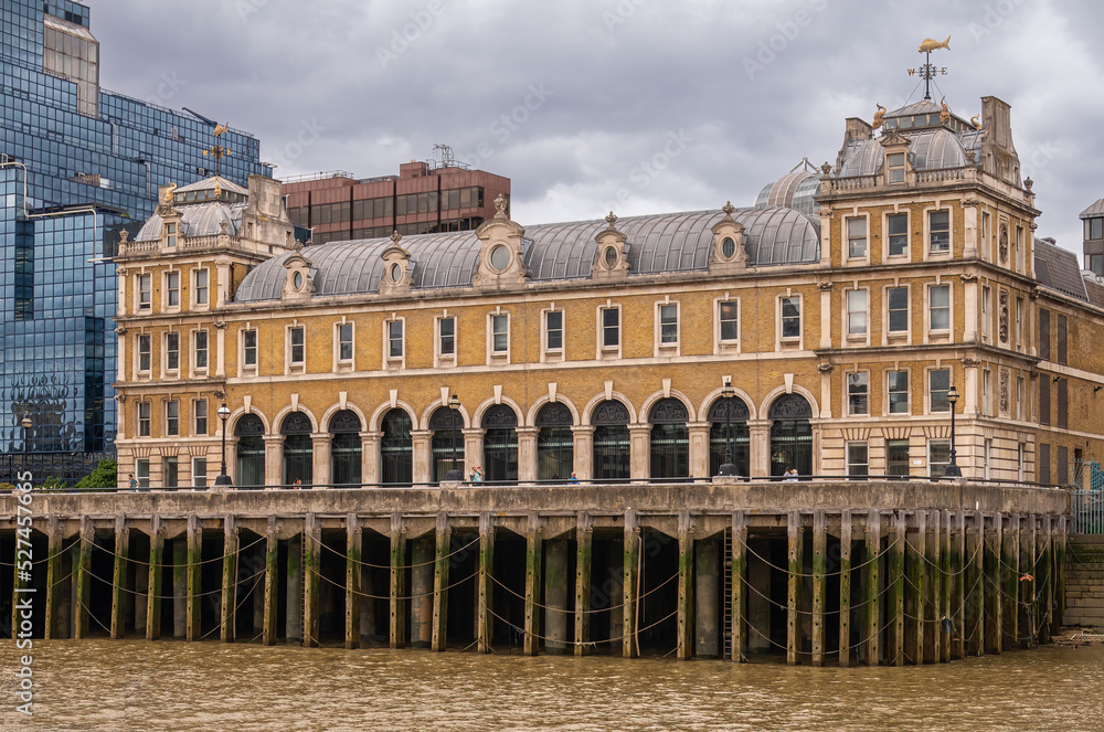 London, England, UK - July 6, 2022: From Thames River. North shore, Old Billingsgate historic yellow stones building with corner towers and gold fishes as decoration on top. under gray sky.