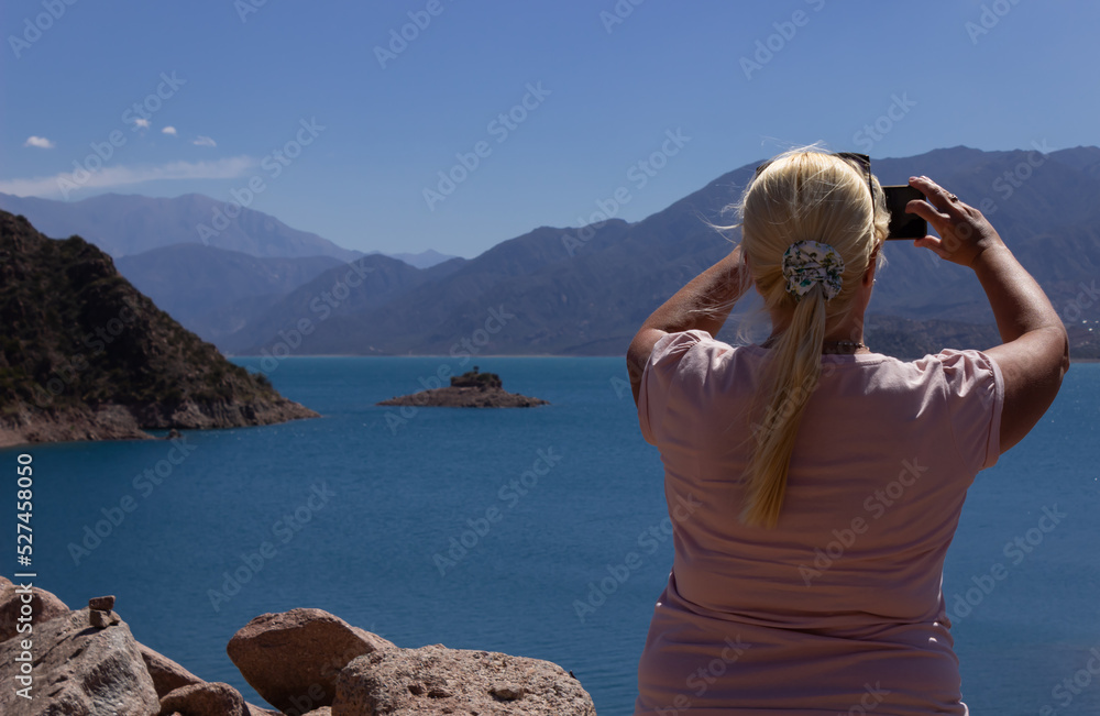 Blonde woman taking a picture in a beautiful scenery with a lake and mountains on a cloudy day.