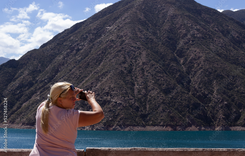 Blonde woman taking a picture in a beautiful scenery with a lake and mountains on a cloudy day. Blonde woman taking a picture in a beautiful scenery with a lake and mountains on a cloudy day.