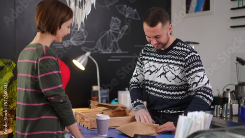 Male cashier packing delicious croissant for client in coffee shop smiling. Portrait of confident Caucasian man placing dessert in paper bag passing purchase to smiling woman in cafe