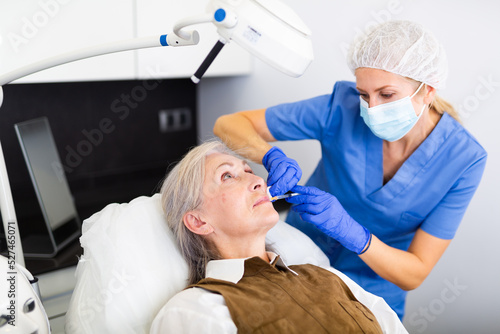 Experienced female cosmetologist working with senior female client in clinic of aesthetic medicine  injecting facial filler into skin to correct face contour