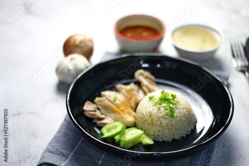 Hainanese chicken rice served on a black plate with dipping sauce and hot broth on white table.