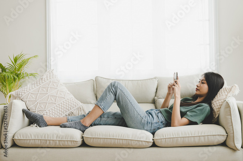 Happy young asian woman relaxing at home she is lying down on sofa and using mobile smartphone