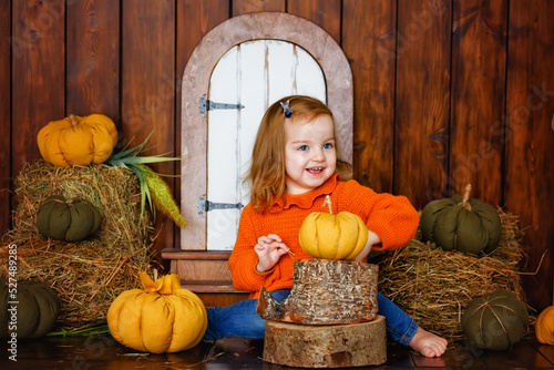 A charming two-year-old girl is sitting in the studio next to a hay on a wooden background and playing with textile pumpkins. The concept of autumn.