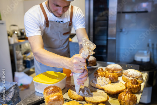 A male baker in a small artisan bakery fills brioches with cream. Front view.