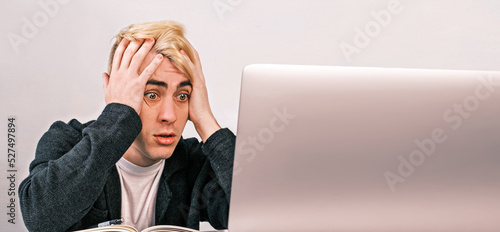 Young office worker is shocked and surprised looking at a laptop clutching his head, sitting at a desk in an office surrounded by documents, notebooks and folders. Success, good deal, unluck