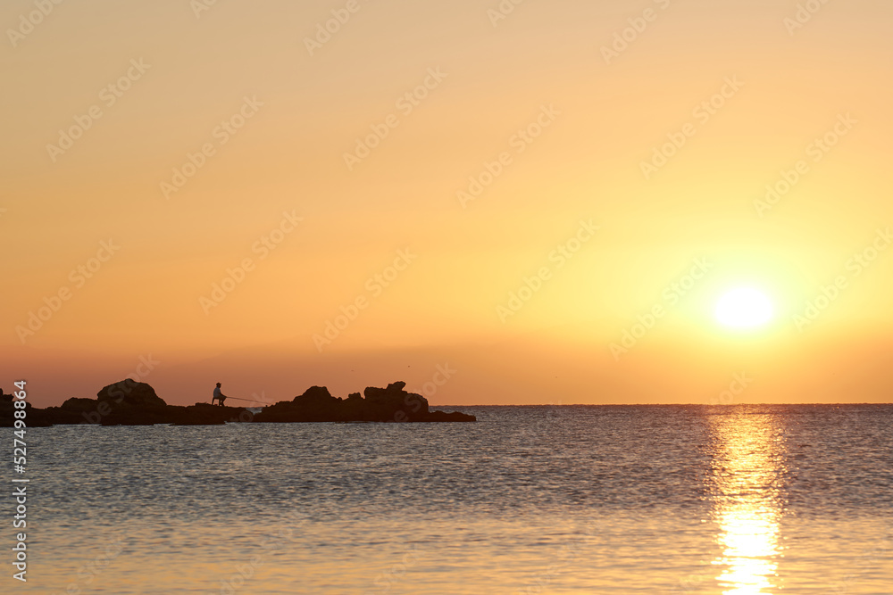 A man is fishing by the mediterranean sea with the rising morning sun in the background