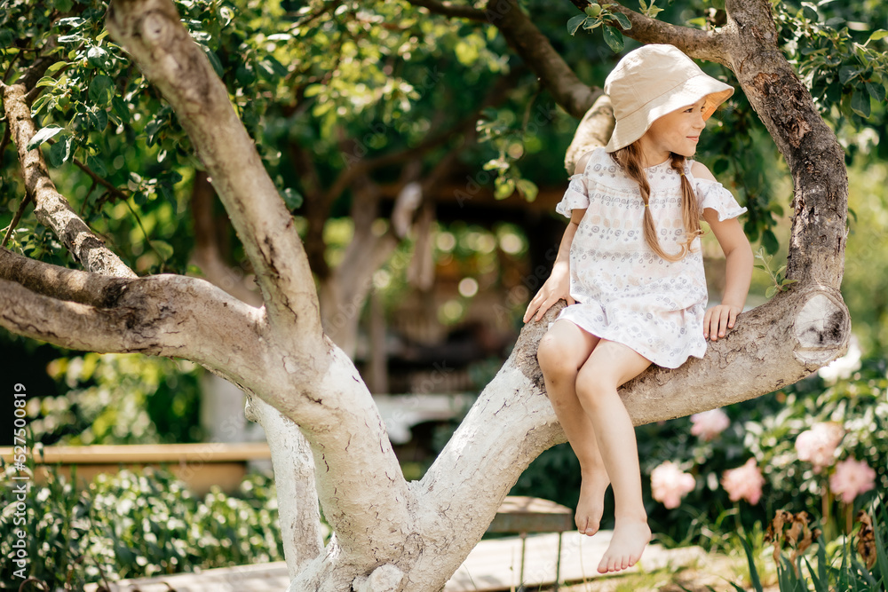 Fototapeta premium Small girl climb tree in summer garden, activity. Little girl on tree branch, childhood. Vacation, activity, lifestyle. Kid fashion, style, beauty Childhood youth growth