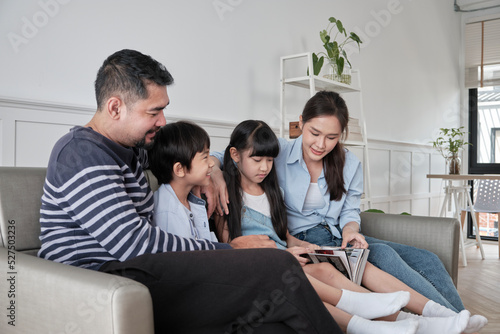 Asian Thai family, adult dad, mum, and children happiness home living relaxing activities and reading book together, leisure on sofa in white room house, lovely weekend, wellbeing domestic lifestyle.