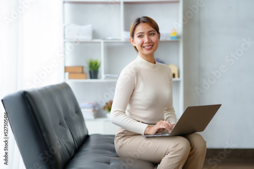 Beautiful Asian businesswoman sitting comfortably on sofa at home with laptop and bright smile of a new day.