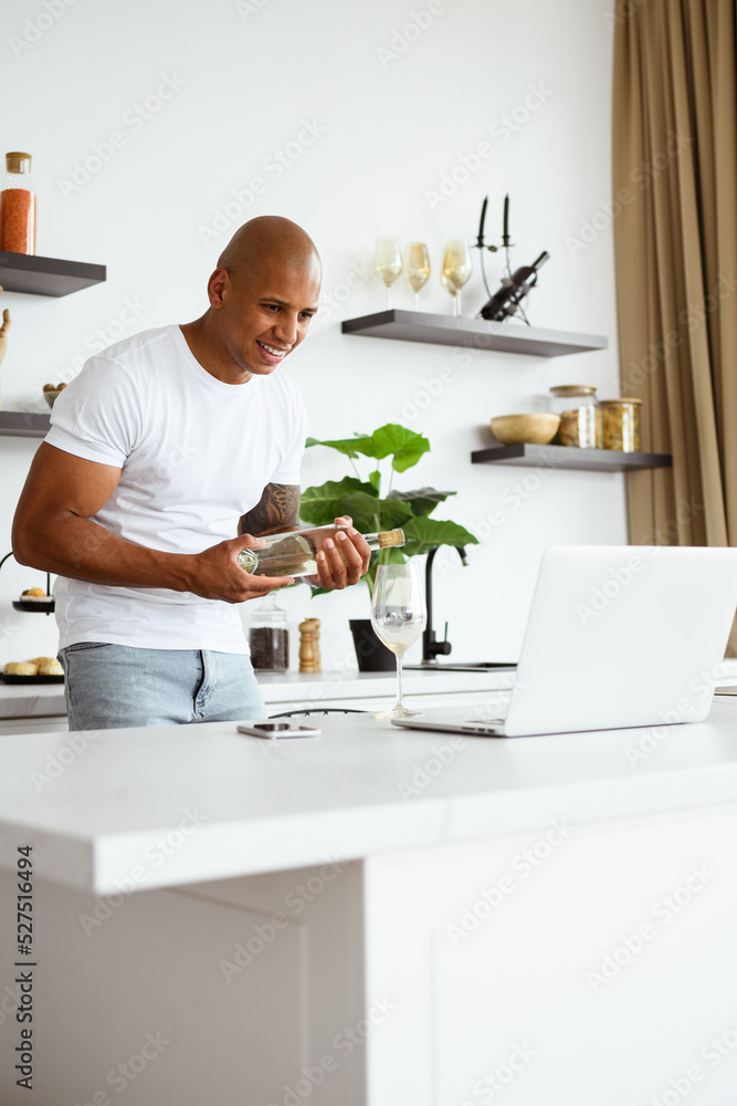 Smiling african american man holding bottle of wine during video call on laptop in kitchen Smiling african american man holding bottle of wine during video call on laptop in kitchen