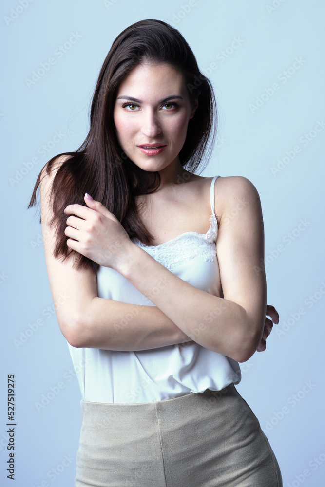 Beautiful woman fashion studio portrait with long dark hair and day make-up with seductive look and pose with crossed arms. Model wearing white blouse and beige trousers. Toned image with blue color Beautiful woman fashion studio portrait with long dark hair and day make-up with seductive look and pose with crossed arms. Model wearing white blouse and beige trousers. Toned image with blue color
