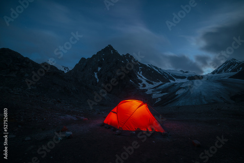 Awesome mountain landscape with vivid orange tent near large glacier tongue under clouds in night starry sky. Tent glow by orange light with view to glacier and mountains silhouettes in starry night. photo