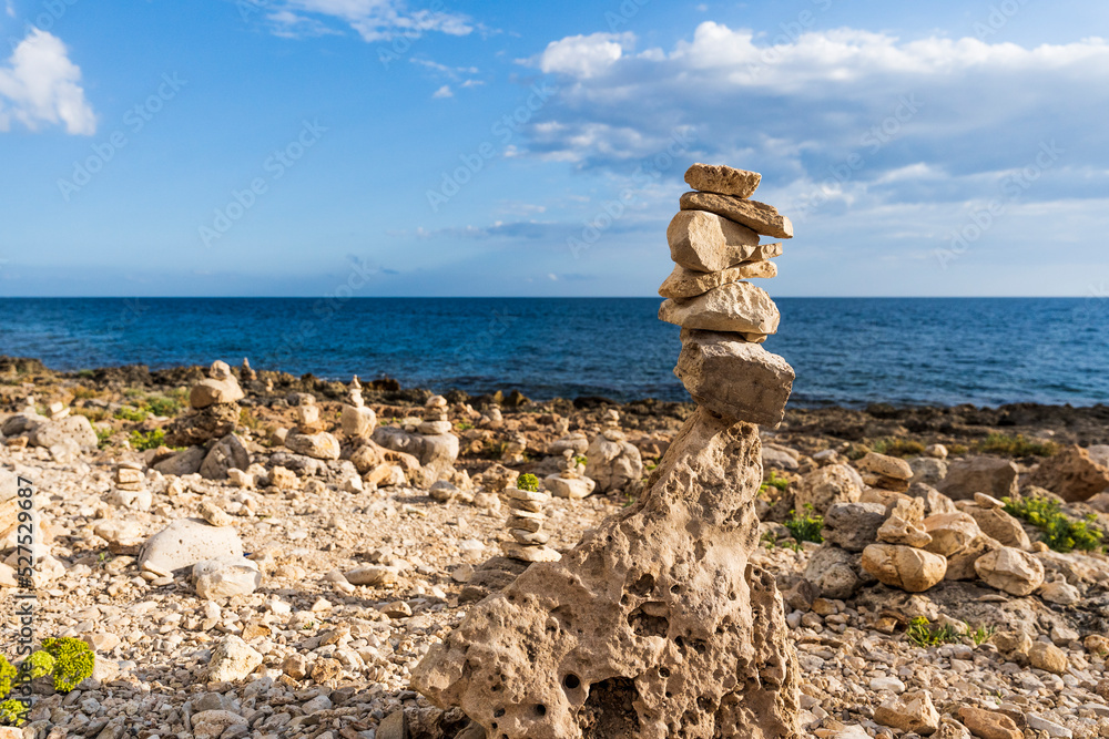 A man-shaped cairn made of rocks along the shore of Capilungo, Salento, Puglia, Southern Italy, in a sunny summer day, with bright colors, clear water and blue sky. A man-shaped cairn made of rocks along the shore of Capilungo, Salento, Puglia, Southern Italy, in a sunny summer day, with bright colors, clear water and blue sky.