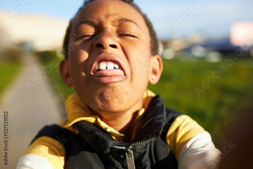 Fotografie, Obraz Funny self-portrait of african american boy kid grimacing, making faces playing with smartphone camera taking selfie outdoor while walking in park