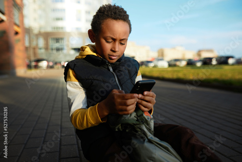 Lost black kid boy dialing number on smartphone to call his parents sitting on pavement in city street or unknown neighborhood, returning home from school, looking nervous and anxious