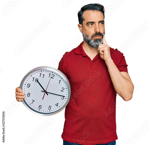 Middle aged man with beard holding big clock serious face thinking about question with hand on chin, thoughtful about confusing idea
