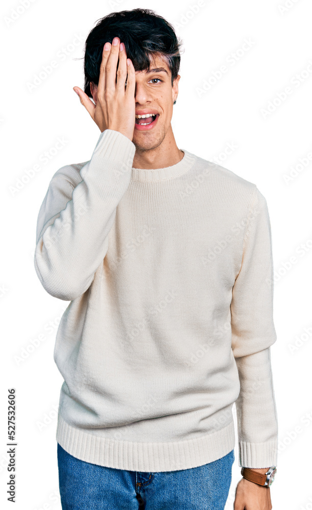Handsome hipster young man wearing casual winter sweater covering one eye with hand, confident smile on face and surprise emotion. Handsome hipster young man wearing casual winter sweater covering one eye with hand, confident smile on face and surprise emotion.
