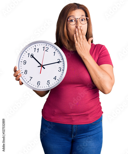 Middle age latin woman holding big clock covering mouth with hand, shocked and afraid for mistake. surprised expression