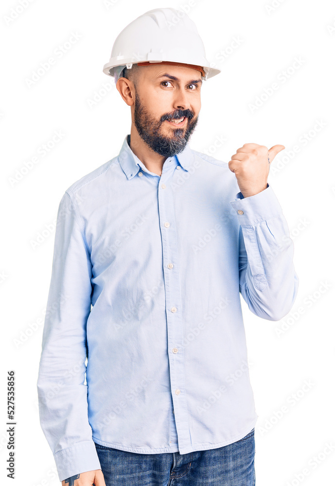 Young handsome man wearing architect hardhat smiling with happy face looking and pointing to the side with thumb up. Young handsome man wearing architect hardhat smiling with happy face looking and pointing to the side with thumb up.