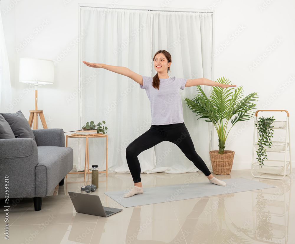 Young woman is taking yoga lesson online on laptop and standing to practicing yoga with pose warrior Young woman is taking yoga lesson online on laptop and standing to practicing yoga with pose warrior