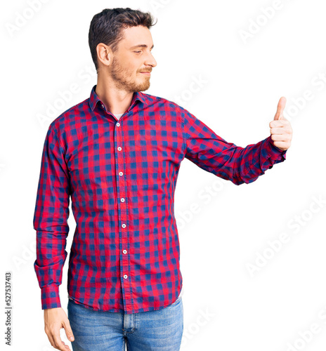 Handsome young man with bear wearing casual shirt looking proud, smiling doing thumbs up gesture to the side