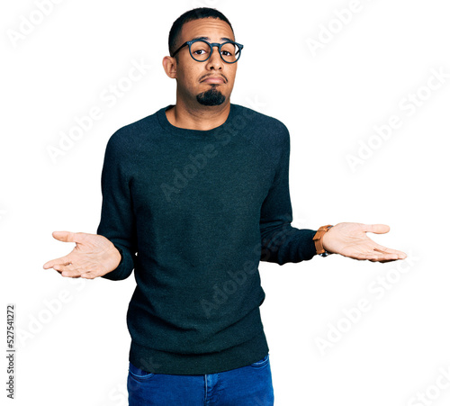 Принти на полотні Young african american man wearing casual clothes and glasses clueless and confused expression with arms and hands raised