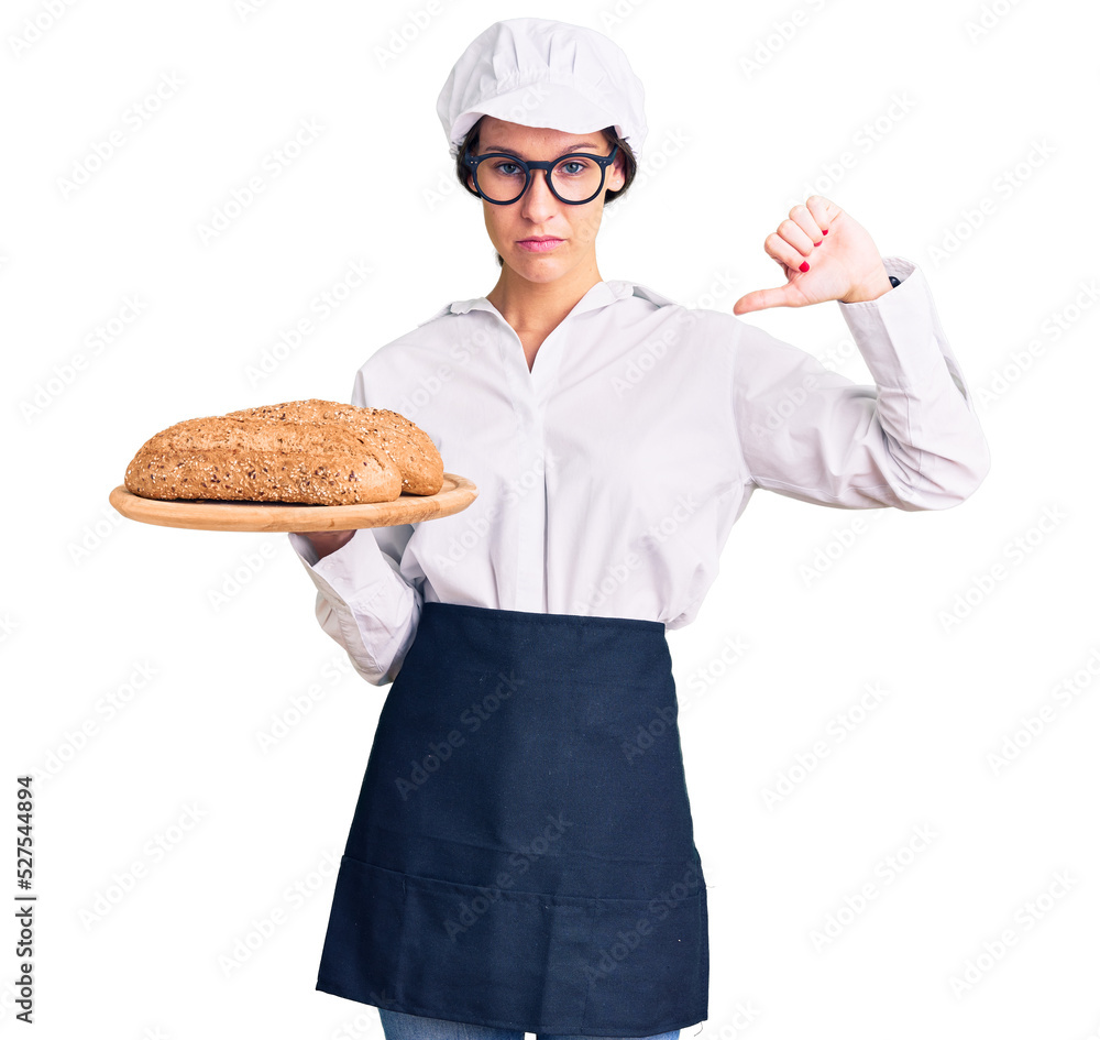 Beautiful brunette young woman wearing baker uniform holding homemade bread with angry face, negative sign showing dislike with thumbs down, rejection concept Beautiful brunette young woman wearing baker uniform holding homemade bread with angry face, negative sign showing dislike with thumbs down, rejection concept