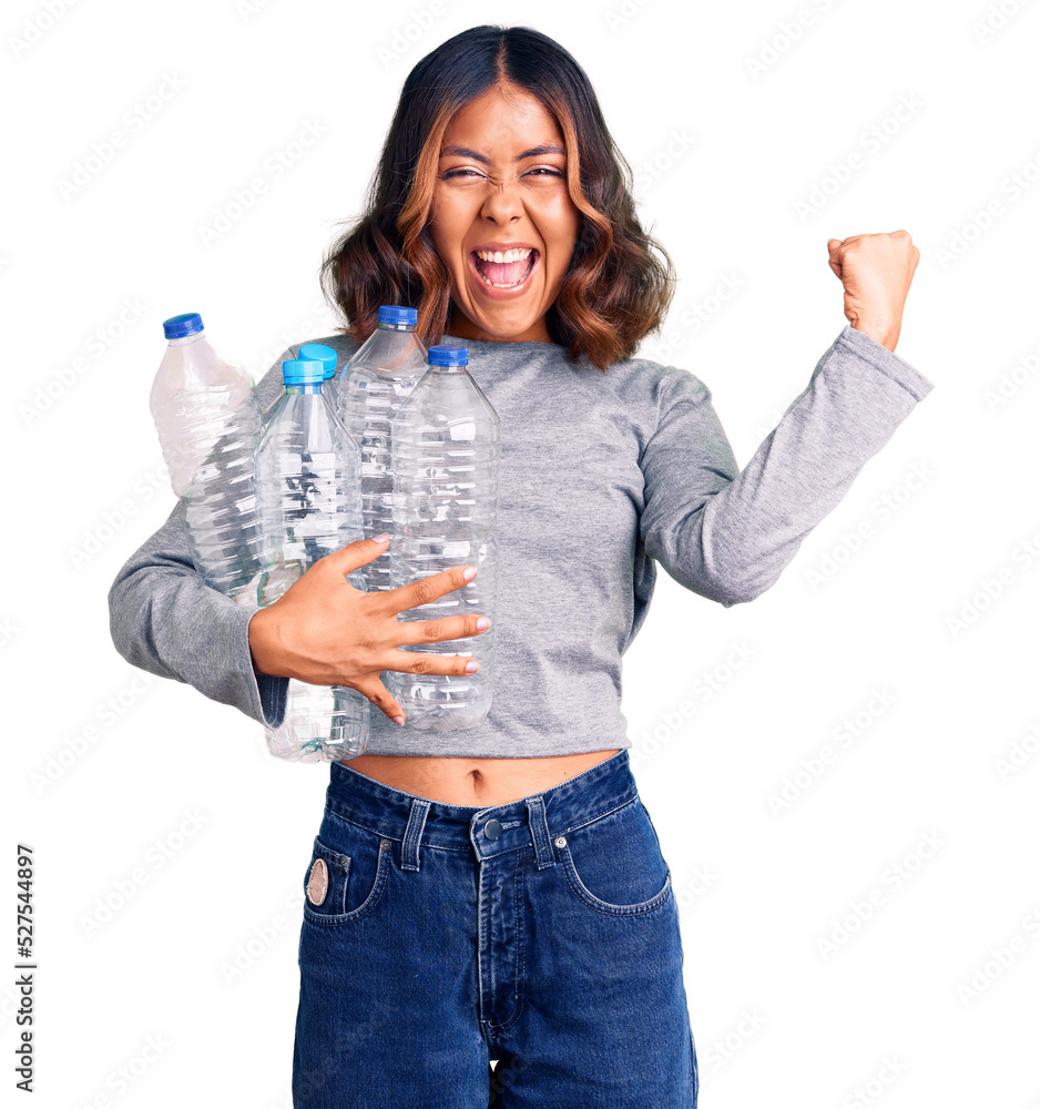 Young beautiful mixed race woman holding recycling plastic bottles screaming proud, celebrating victory and success very excited with raised arms Young beautiful mixed race woman holding recycling plastic bottles screaming proud, celebrating victory and success very excited with raised arms