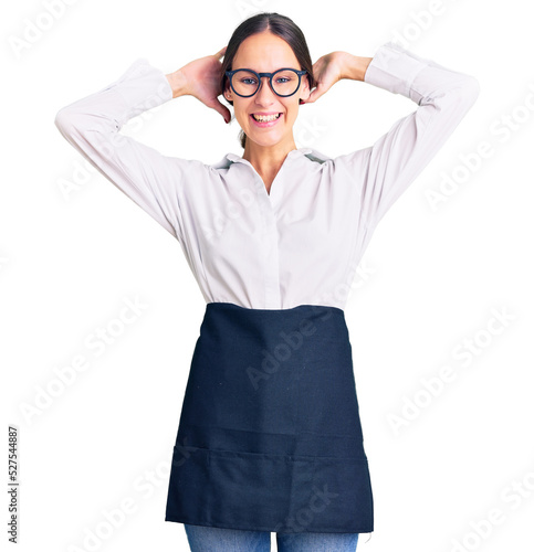 Beautiful brunette young woman wearing professional waitress apron relaxing and stretching, arms and hands behind head and neck smiling happy