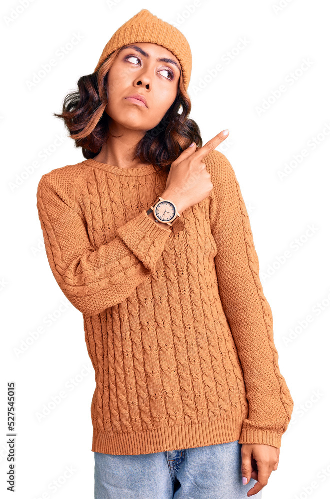 Young beautiful mixed race woman wearing wool sweater and winter hat pointing with hand finger to the side showing advertisement, serious and calm face Young beautiful mixed race woman wearing wool sweater and winter hat pointing with hand finger to the side showing advertisement, serious and calm face