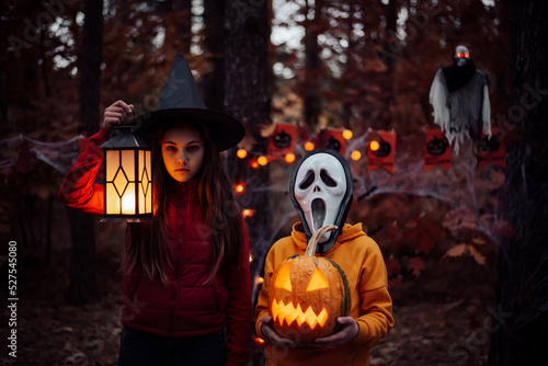 Two children in halloween costumes in the woods. Autumn holiday decoration, jack-o-lantern, creepy mask, witch hat, ghost on the background.