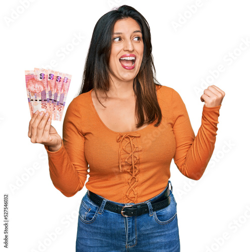 Beautiful hispanic woman holding south african 50 rand banknotes pointing thumb up to the side smiling happy with open mouth