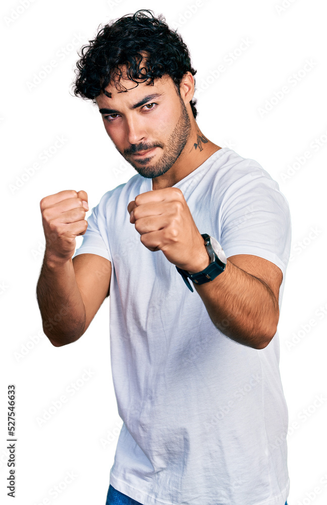 Hispanic young man with beard wearing casual white t shirt ready to fight with fist defense gesture, angry and upset face, afraid of problem Hispanic young man with beard wearing casual white t shirt ready to fight with fist defense gesture, angry and upset face, afraid of problem