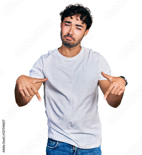 Hispanic young man with beard wearing casual white t shirt pointing down looking sad and upset, indicating direction with fingers, unhappy and depressed.