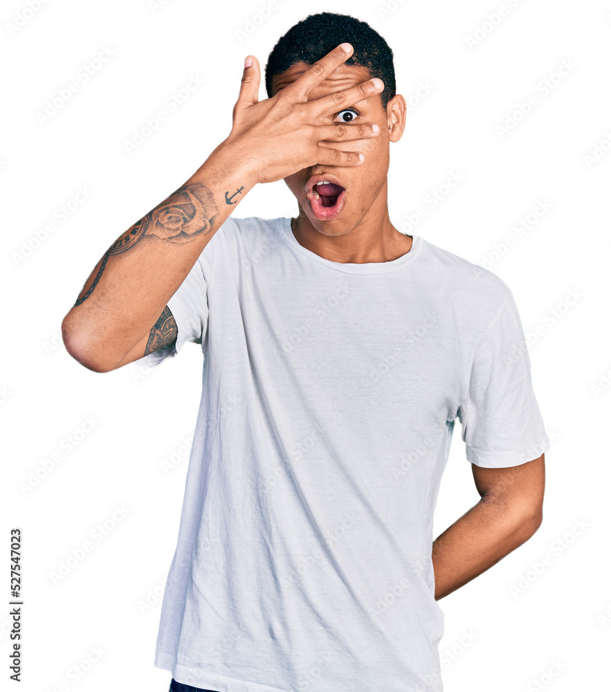 Young hispanic man wearing casual white t shirt peeking in shock covering face and eyes with hand, looking through fingers with embarrassed expression. Young hispanic man wearing casual white t shirt peeking in shock covering face and eyes with hand, looking through fingers with embarrassed expression.