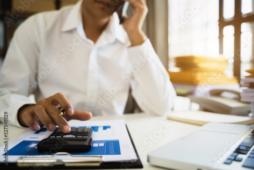 Male businessman working on desk office with using a calculator to calculate the numbers and business report talk smart phone on office desk  finance accounting concept.