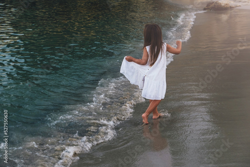 A little girl in a white dress walks along the line of the sea. photo