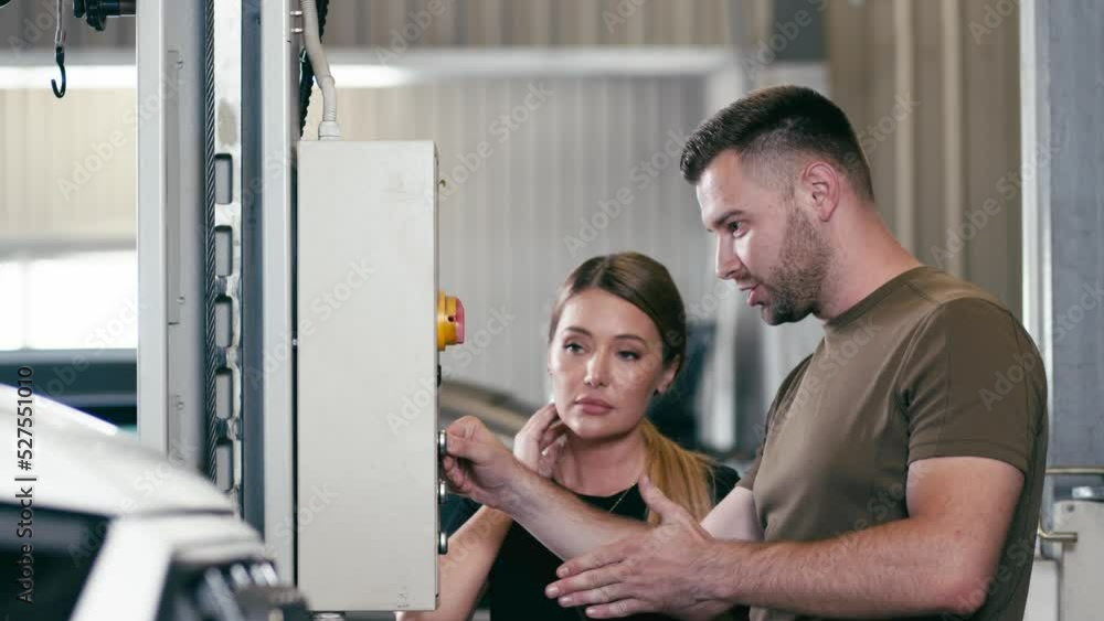 A woman mechanic prepares a car for diagnostics on a special lift and explains the breakdown of the car to the manager. Car service workers talking while lifting the car in the garage. A woman mechanic prepares a car for diagnostics on a special lift and explains the breakdown of the car to the manager. Car service workers talking while lifting the car in the garage.