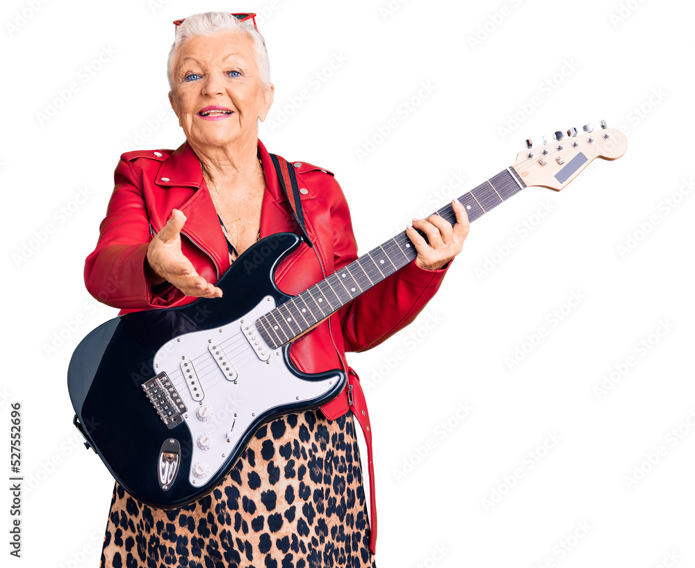 Senior beautiful woman with blue eyes and grey hair wearing a modern look playing electric guitar smiling cheerful offering palm hand giving assistance and acceptance.