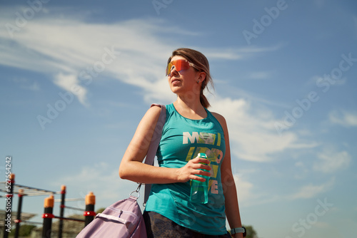 Female athlete with violet bag on her shoulder and water bottle stands near stadium waiting for training.