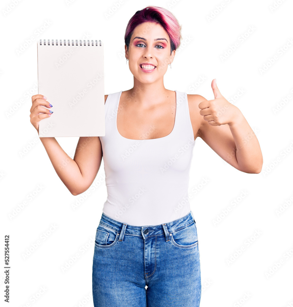 Young beautiful woman with pink hair holding notebook smiling happy and positive, thumb up doing excellent and approval sign Young beautiful woman with pink hair holding notebook smiling happy and positive, thumb up doing excellent and approval sign