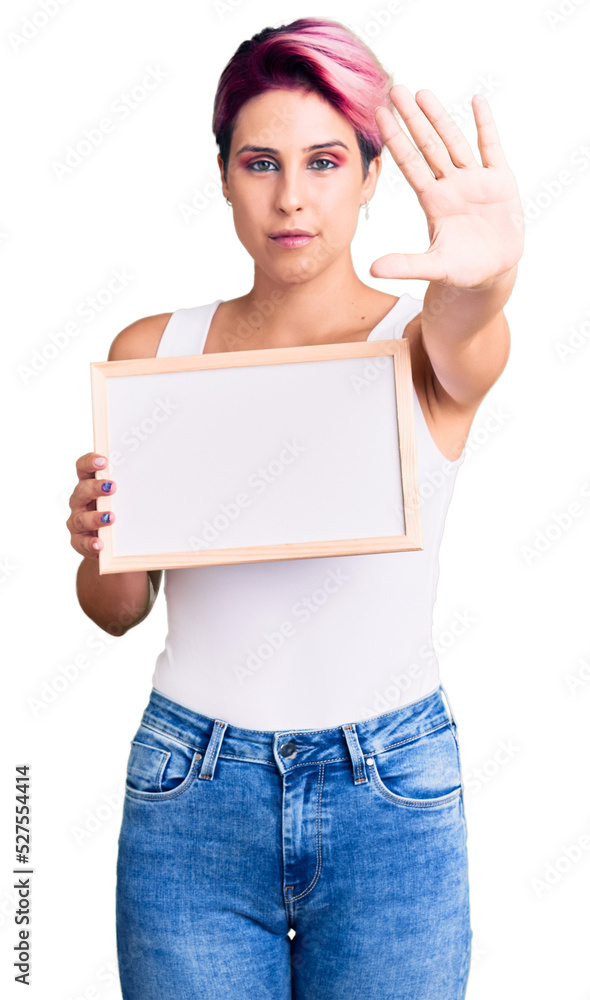 Young beautiful woman with pink hair holding empty white chalkboard with open hand doing stop sign with serious and confident expression, defense gesture Young beautiful woman with pink hair holding empty white chalkboard with open hand doing stop sign with serious and confident expression, defense gesture