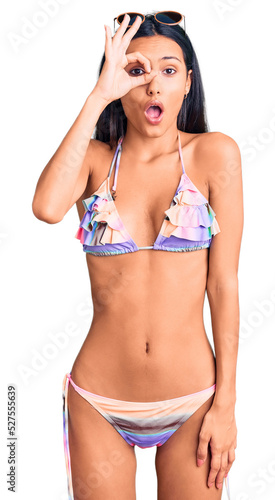Young beautiful latin girl wearing bikini and sunglasses doing ok gesture shocked with surprised face, eye looking through fingers. unbelieving expression.