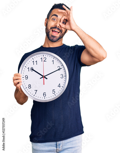 Young hispanic man holding big clock smiling happy doing ok sign with hand on eye looking through fingers